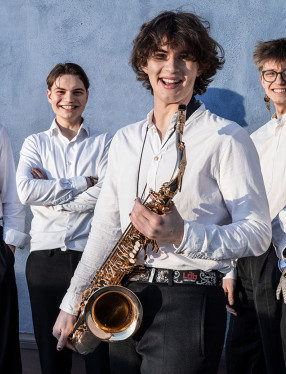 Fem unga musiker, vissa med instrument i handen, ler mot kameran. Fotografi.